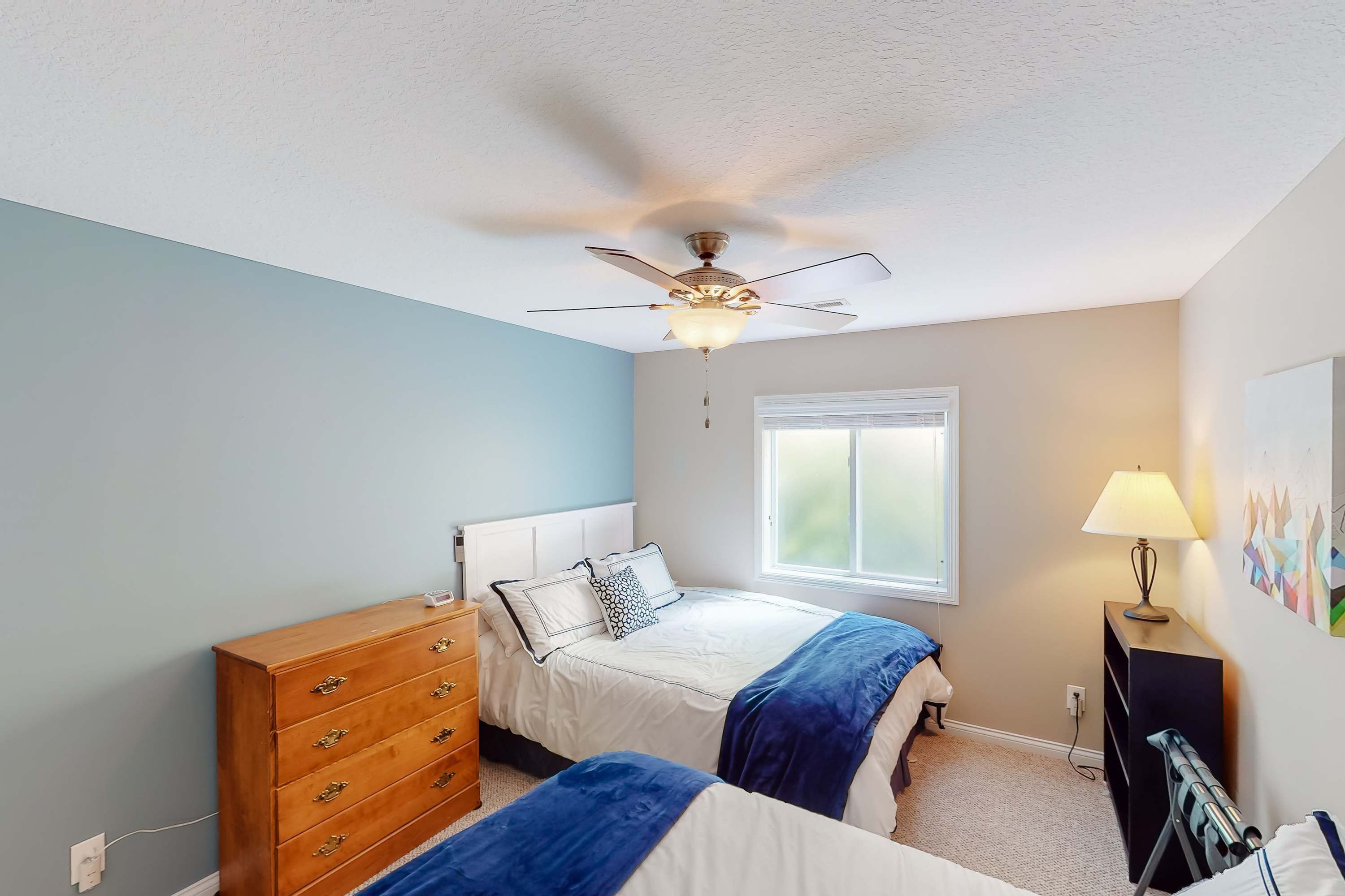 A bedroom with a ceiling fan, a dresser, two beds with blue and white bedding, a window with blinds, and a lamp on a side table.