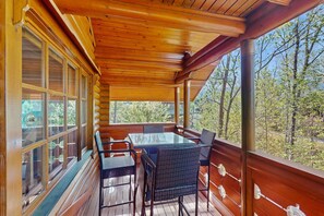 Un terrazzo in legno con tavolo e sedie in vetro, circondato da ampie finestre che si affacciano su alberi.