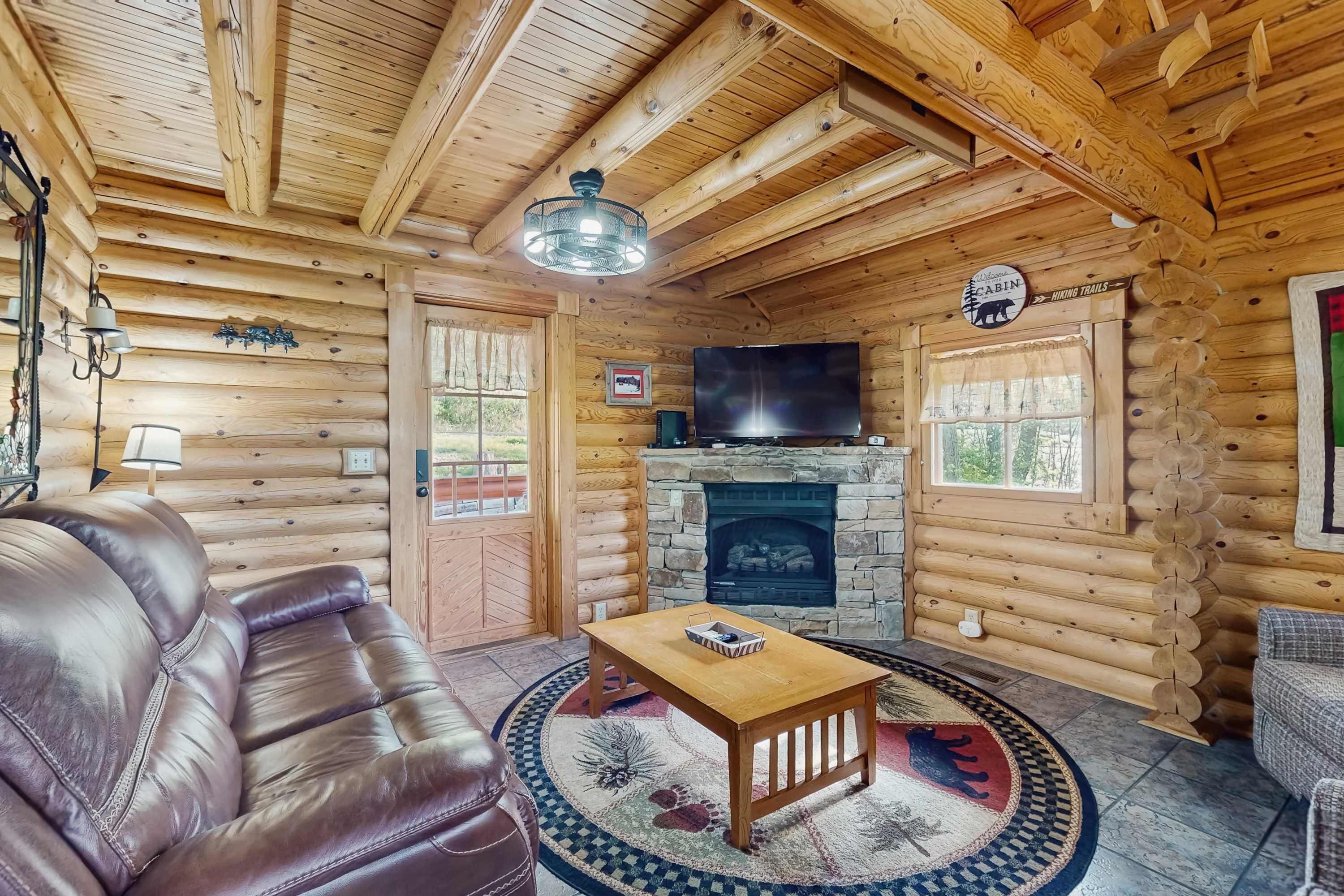 Interior de una cabaña de troncos con un sofá de cuero, una mesa de centro de madera, una chimenea, un televisor y un letrero que indica cabañas y senderos para caminatas.