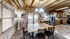Interior de uma cabana de madeira com uma mesa de jantar, cadeiras, um lustre e uma cozinha.