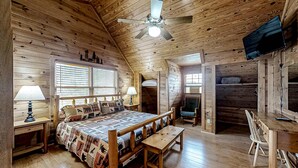 Quarto de cabana de madeira com uma cama, duas mesas de cabeceira com abajures, um banco de madeira, uma escrivaninha e uma televisão fixada na parede.