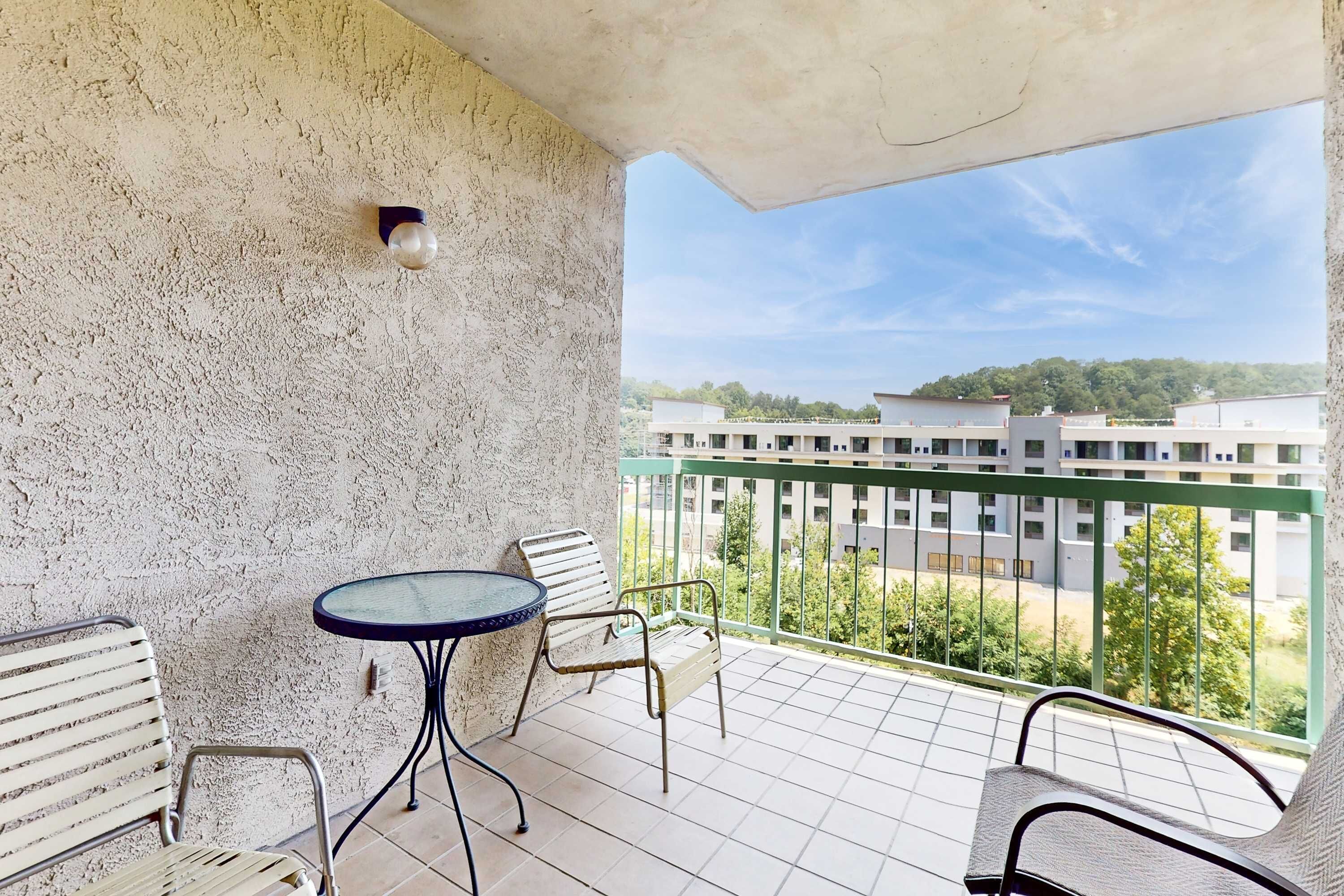 A balcony with a table and chairs, a textured wall, and a view of buildings and greenery.