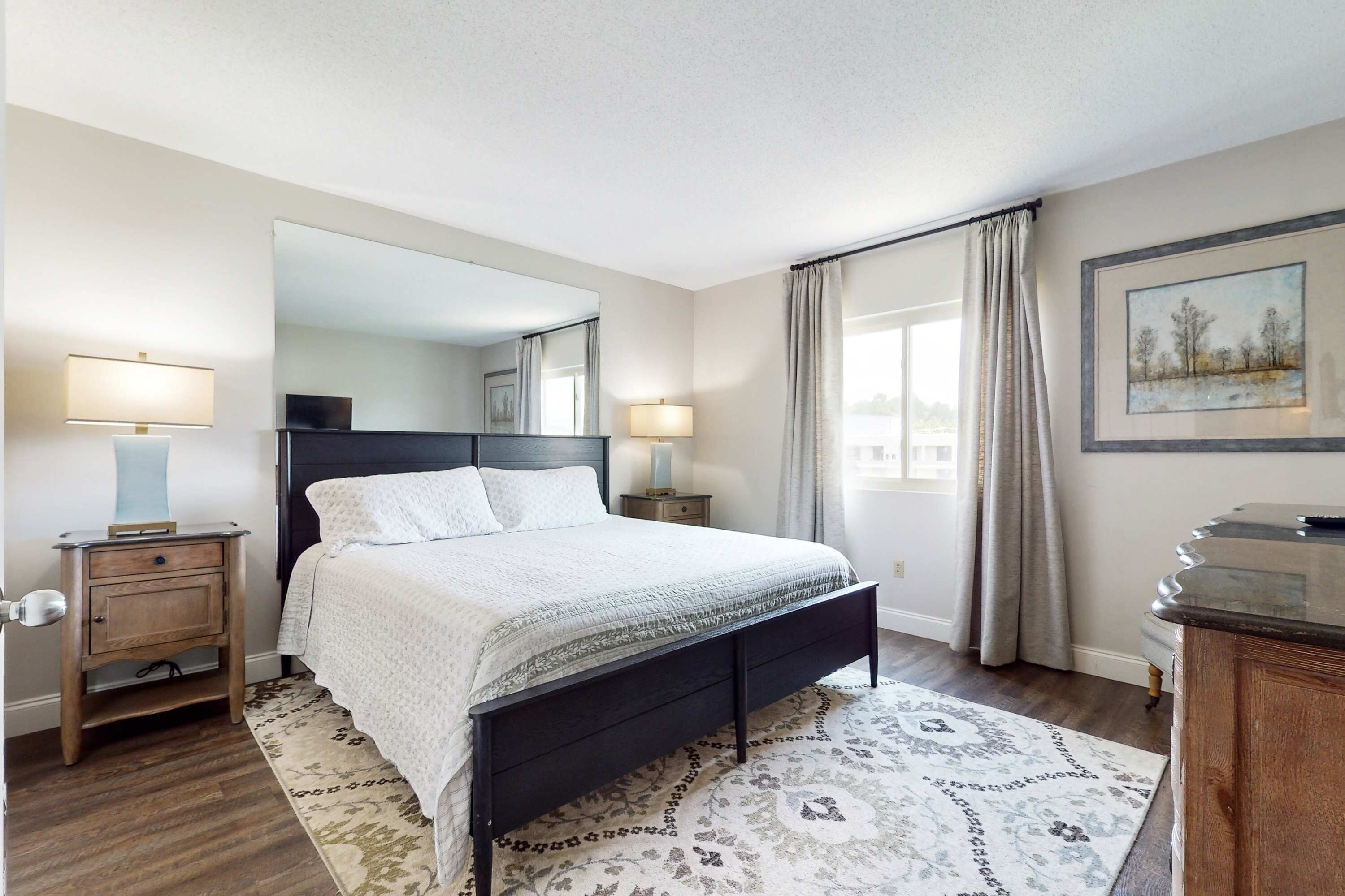 Quarto com cama, mesas de cabeceira, espelho, cômoda e janela com cortinas.