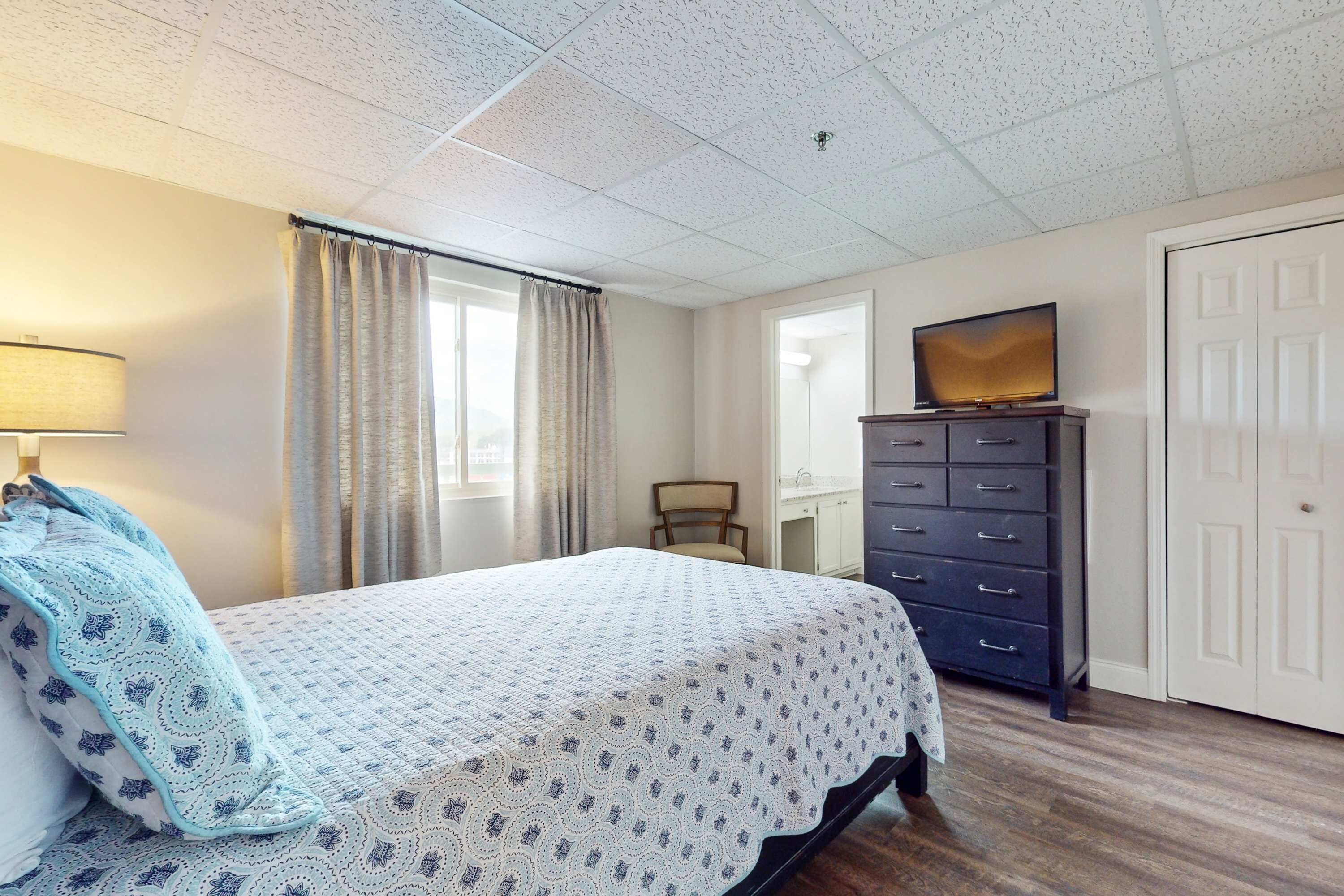 Une chambre à coucher avec un lit, une commode, une chaise, un téléviseur et une porte.