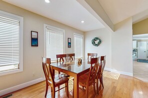 A dining area with a glass-top table surrounded by wooden chairs, large windows with blinds, and a kitchen visible in the background. - Lovely Norris Lakefront Home・Firepit・Dock・Grill (Maynardville)
