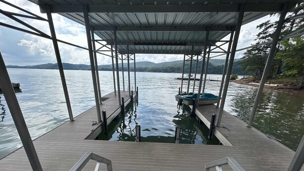 Une jetée en bois avec des garde-corps en métal, un espace abrité et une vue sur un lac paisible, avec des bateaux et des collines au loin.