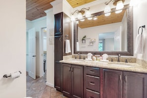 A bathroom with dark wood cabinets, a granite countertop, a large mirror with lights, and a door leading to another room. - Lovely Norris Lakefront Home・Firepit・Dock・Grill (Maynardville)