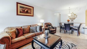 A living room with a brown leather sofa, a coffee table, a dining table with chairs, a chandelier, and a framed picture on the wall. - Cozy Riverfront Downtown Condo, Sleeps 6, Balcony (Pigeon Forge)