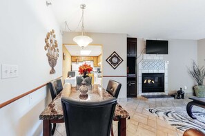 Un comedor con una mesa de mármol, sillas negras y un jarrón de flores. Hay una chimenea con un televisor montado encima.