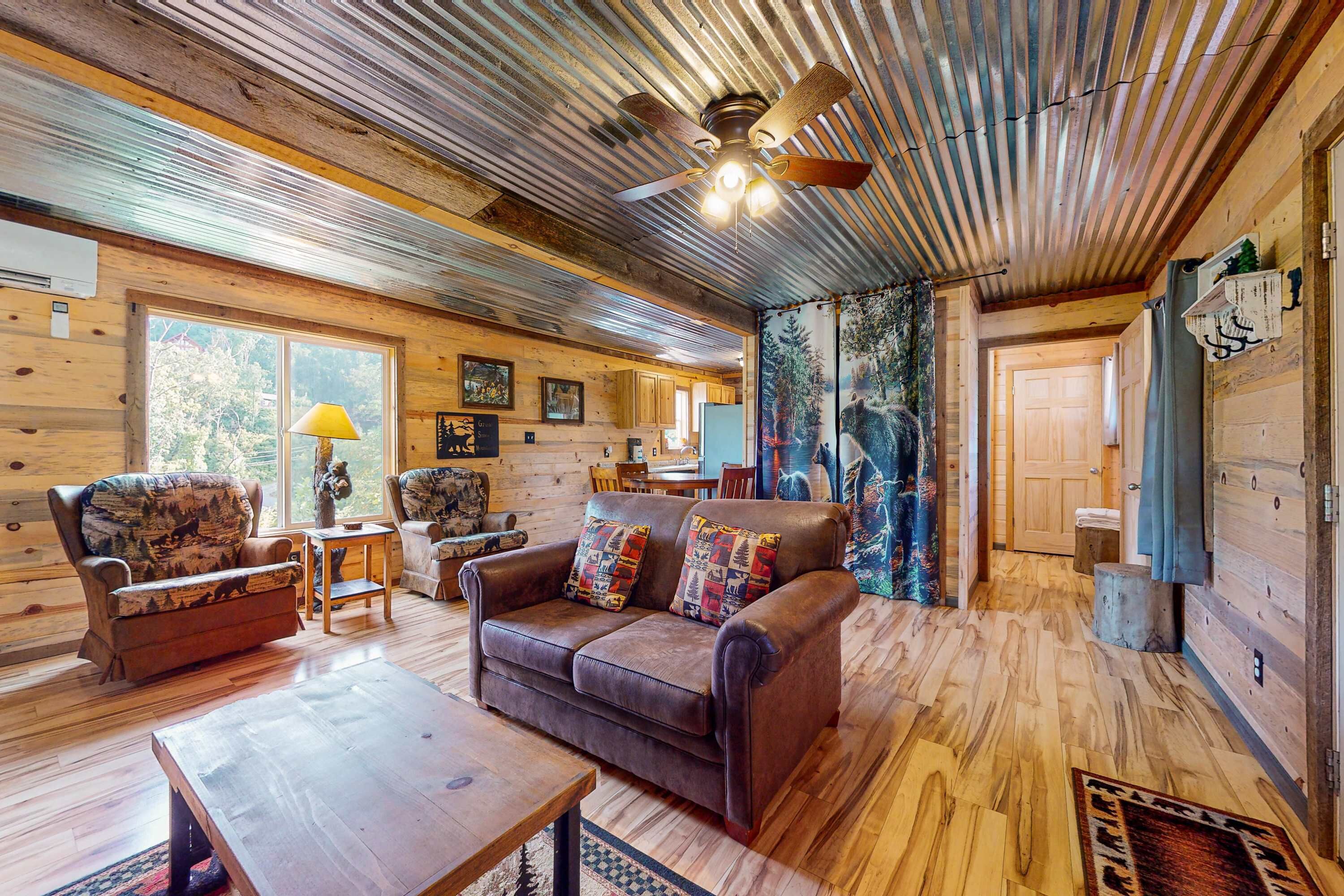 A cozy cabin interior with a wooden floor, leather sofas, a ceiling fan, and a large window.