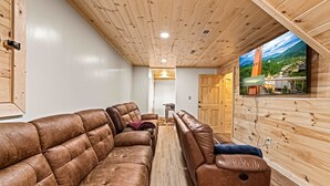 A room with a wooden ceiling, a brown leather sofa, a wooden bench, and a TV screen displaying a scenic view. - Cabin Views, Sleeps 12・Foosball・Theater・Putt-putt (Sevierville)