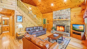 Interior de una cabaña de madera con chimenea de piedra, sofá de cuero y televisor.