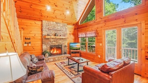 Interior de una cabaña de madera con chimenea de piedra, muebles de cuero y un televisor.