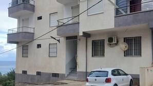 Un immeuble résidentiel à plusieurs étages, doté de balcons, avec une voiture garée devant et une antenne parabolique sur le côté.