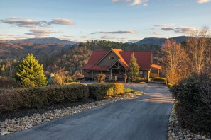 A large, two-story house with a red roof, surrounded by a stone pathway and lush greenery, set against a backdrop of mountains and a clear sky. - Mtn Getaway, Sleeps 12・Spectacular Views・Hot Tub (Sevierville)