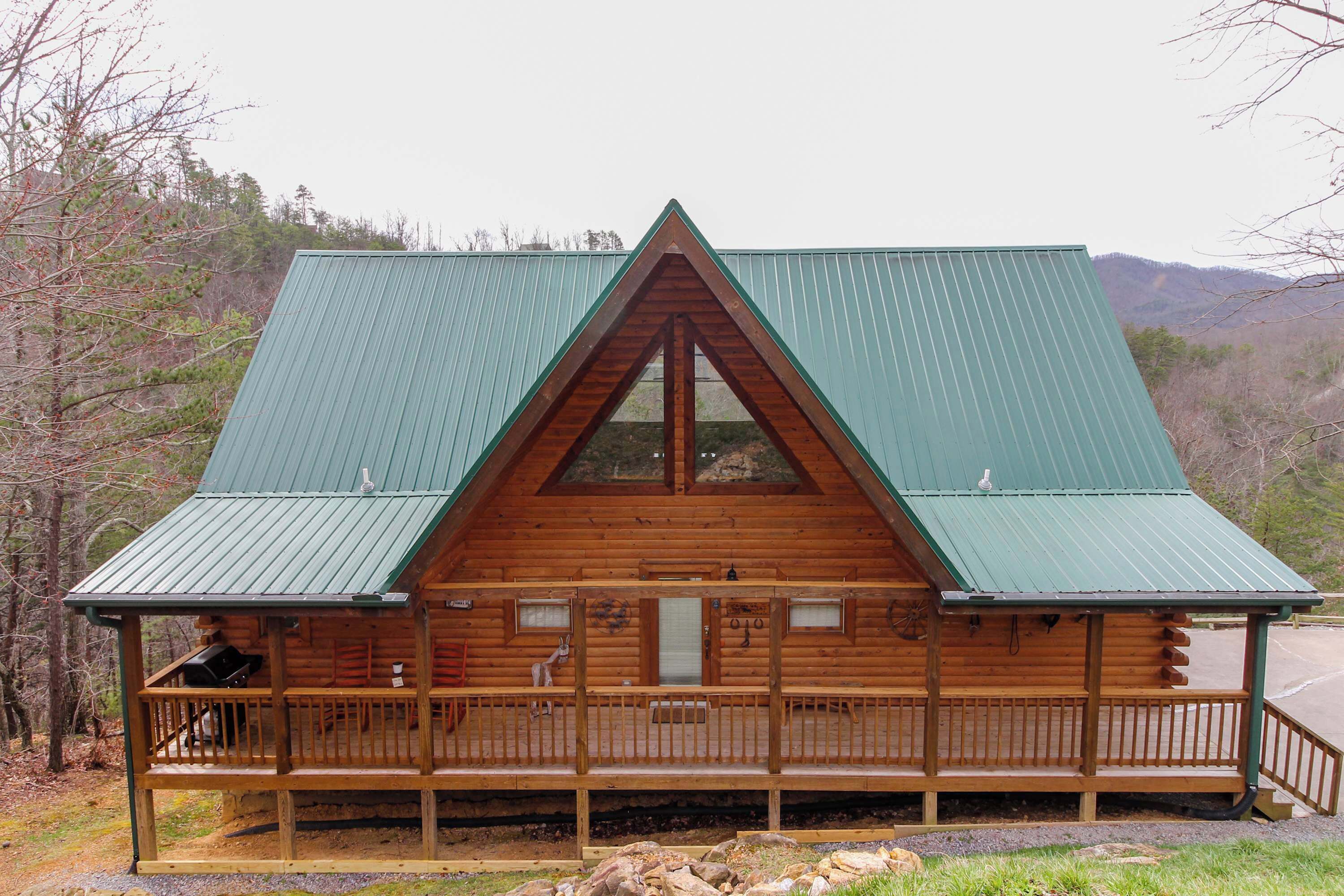 A wooden cabin with a green roof, a porch, and a view of the surrounding forest.