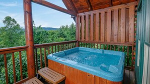 Une terrasse en bois avec un jacuzzi, des bancs en bois et une vue sur des arbres et des collines verdoyantes.