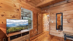 Interior de una cabaña de madera con un televisor sobre un mueble de madera, una ventana con persianas y un espejo en la pared.