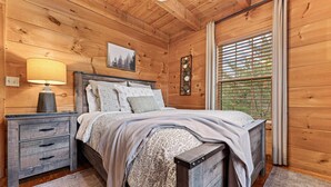 Un dormitorio tipo cabaña de madera con cama, mesita de noche, lámpara y ventana con persianas.