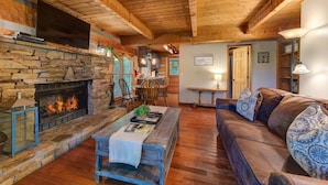 A cozy living room with a stone fireplace, wooden ceiling beams, a leather sofa, and a dining area with wooden chairs and a table. - Dog Friendly Cabin, Sleeps 2・Screened Deck・Jet Tub (Sevierville)