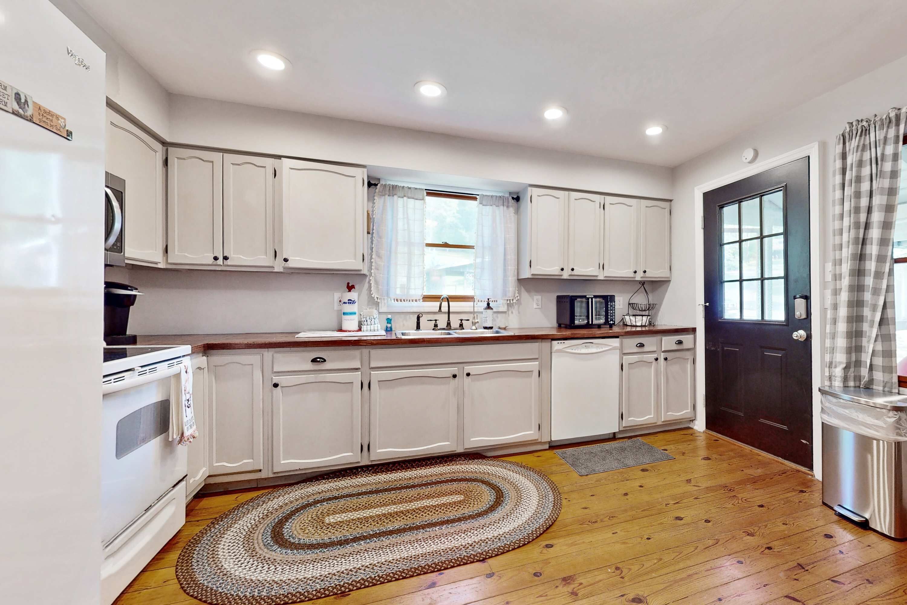 Una cocina con armarios blancos, encimera de madera, horno empotrado, un lavabo y una ventana con cortinas.