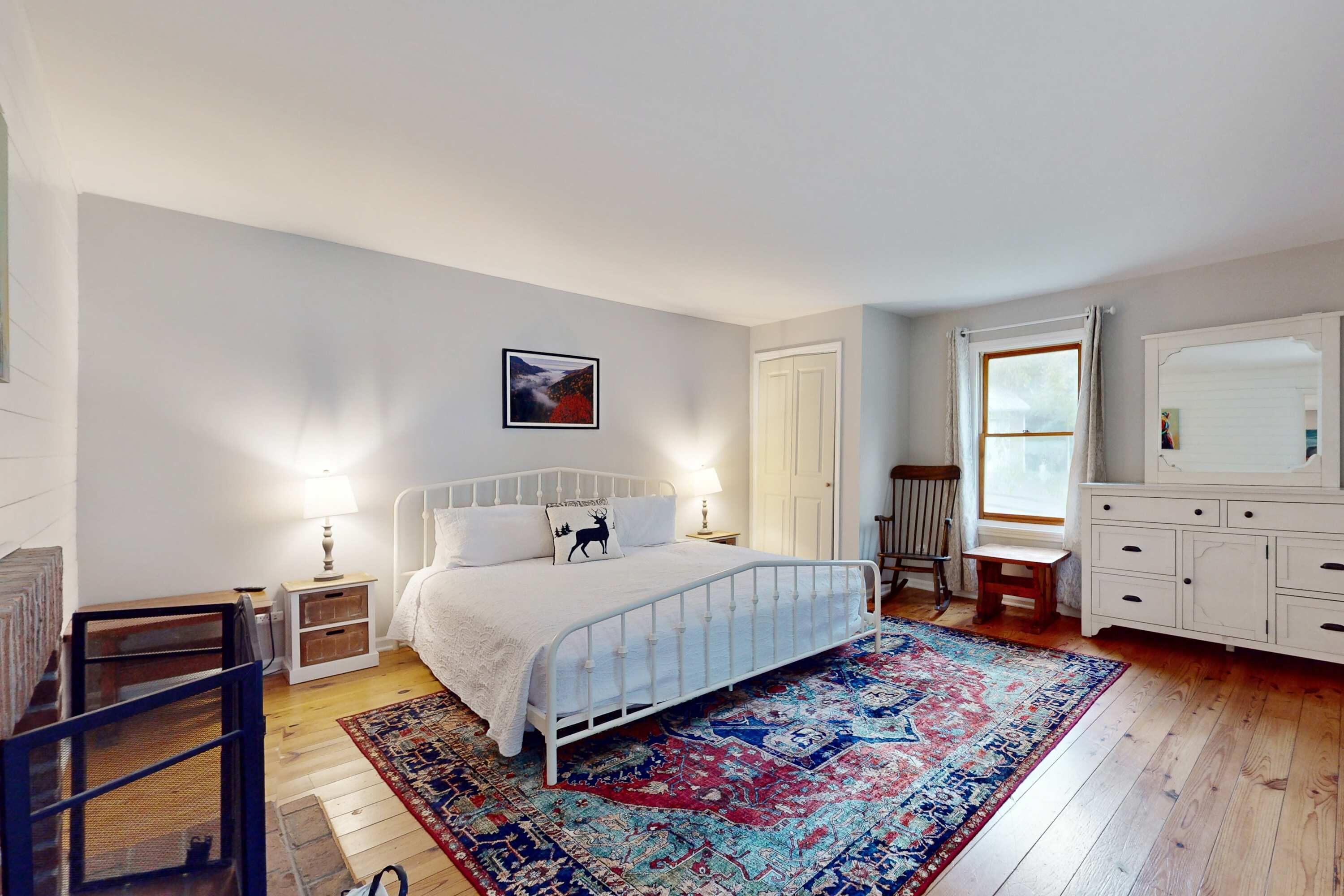 Une chambre avec un lit en métal blanc, un grand tapis à motifs, une chaise en bois, une commode blanche et un tableau encadré accroché au mur.