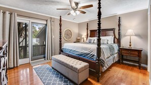 Une chambre à coucher avec un lit à baldaquin, un ventilateur de plafond, un banc, une table de chevet et une vue sur l’extérieur, accessible par une porte-fenêtre coulissante.