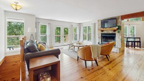 Ein geräumiges Wohnzimmer mit Kamin, Holzboden, einer Couch, einem Esstisch und großen Fenstern mit Blick auf Grünflächen.