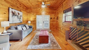 Ein gemütliches Blockhaushaus-Interieur mit Holzbalkendecke, einem grauen Sofa, einem roten Hocker und einem großen Flachbildfernseher.