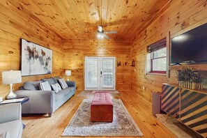 A cozy cabin interior with a wooden ceiling, a gray sofa, a red ottoman, and a large flat-screen TV. - Chalet W Arcade・Pet Ok・Large Yard・Resort Amenities (Sevierville)