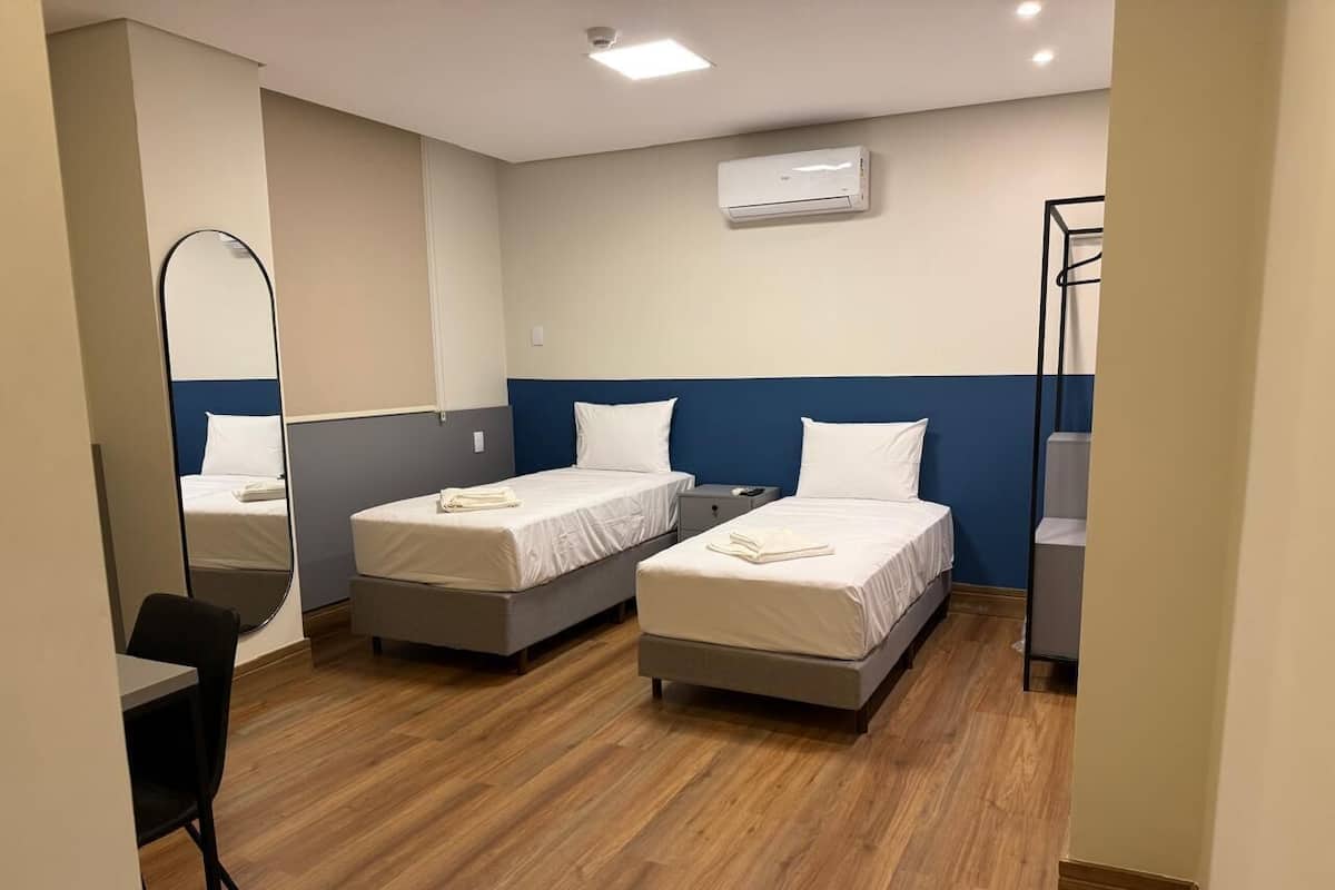 A hotel room with two single beds, a desk, a chair, a mirror, and an air conditioning unit.