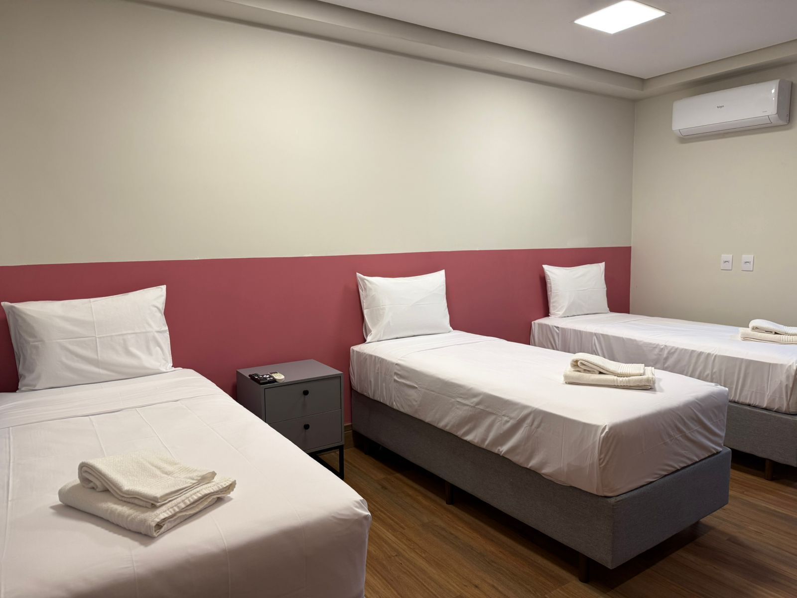 A hotel room with three beds, a bedside table with a phone, and an air conditioning unit on the wall.