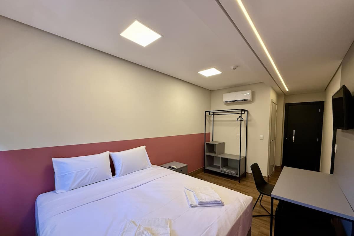 A hotel room with a bed, a desk, a chair, a wardrobe, and an air conditioning unit.