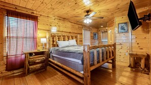 Ein Schlafzimmer einer Holzhütte mit Bett, Nachttisch, Lampe, Deckenventilator und einem an der Wand befestigten Fernseher.