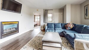 A living room with a blue sofa, a flat-screen TV mounted on the wall, and a fireplace with a decorative frame. - 2 All-Suite Condos, Sleeps & River View Balconies (Pigeon Forge)
