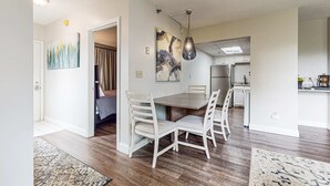 A dining area with a wooden table and white chairs, a kitchen in the background, and artwork on the walls. - 2 All-Suite Condos, Sleeps & River View Balconies (Pigeon Forge)