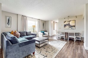 A modern living room with a grey sofa, a coffee table, and a dining area with white chairs and a table. - 2 All-Suite Condos, Sleeps & River View Balconies (Pigeon Forge)