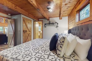 A bedroom with a large bed, wooden ceiling beams, a ceiling fan, and a window with a stained glass design. - Serene Cabin, Sleeps 4・Screened Porch・Games・Grill (Sevierville)