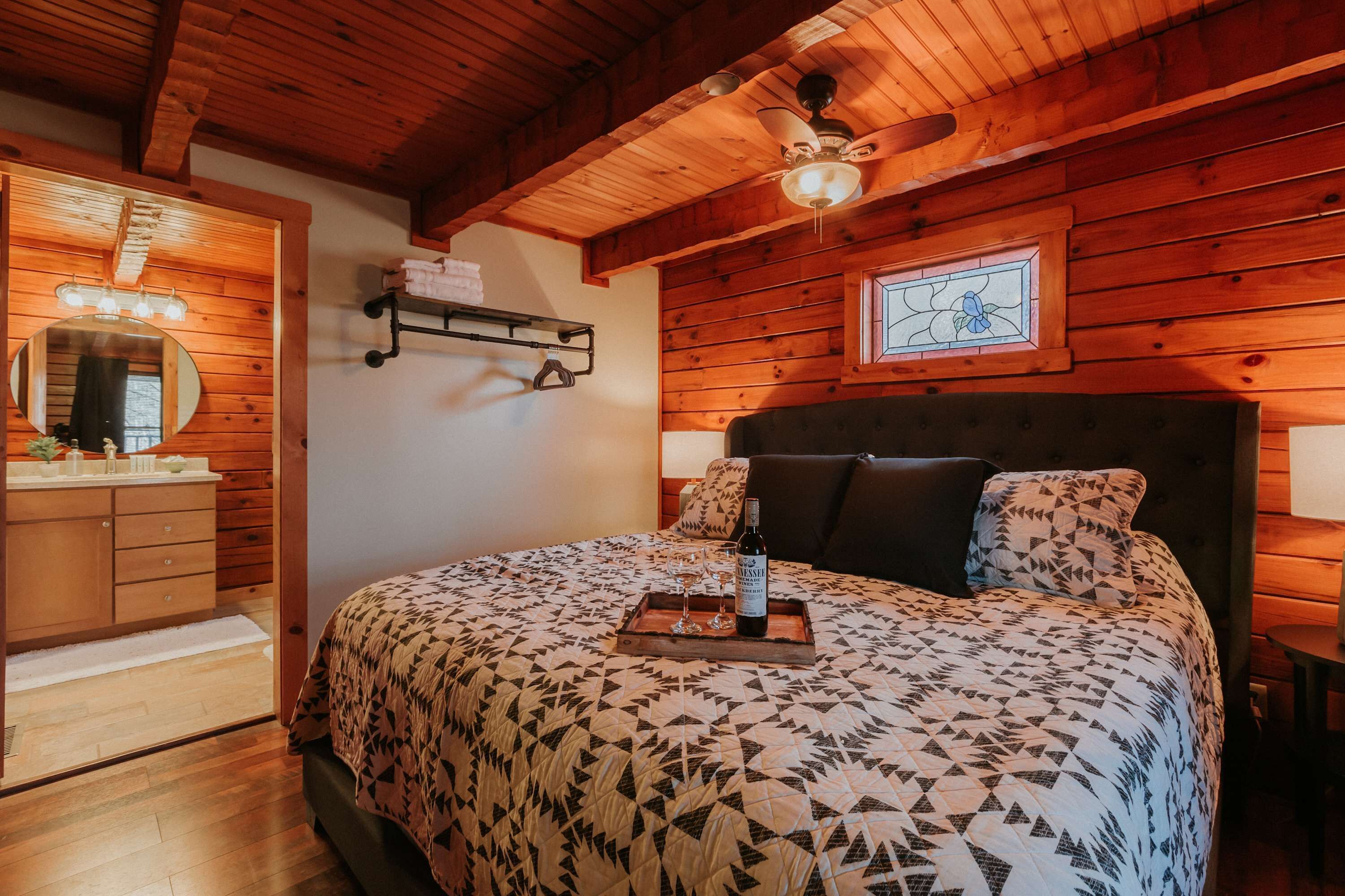 A wooden-paneled bedroom with a bed, a nightstand, a ceiling fan, a mirror, and a window with a stained glass design.