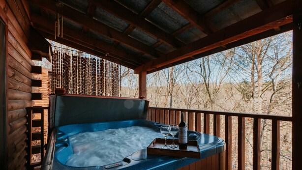 Ein Whirlpool mit Fernbedienung, einer Flasche und zwei Gläsern auf einem Tablett, vor einer Holzterrasse mit Blick auf Bäume.
