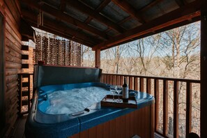 A hot tub with a remote control, a bottle, and two glasses on a tray, set against a wooden deck with a view of trees. - Serene Cabin, Sleeps 4・Screened Porch・Games・Grill (Sevierville)