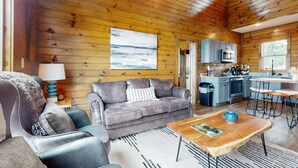 Een gezellig interieur van een chalet met een grijze bank, een houten eettafel en een keuken met kasten en apparatuur.