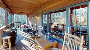 Une terrasse en bois avec une table et des chaises, de grandes fenêtres et une vue sur les arbres.