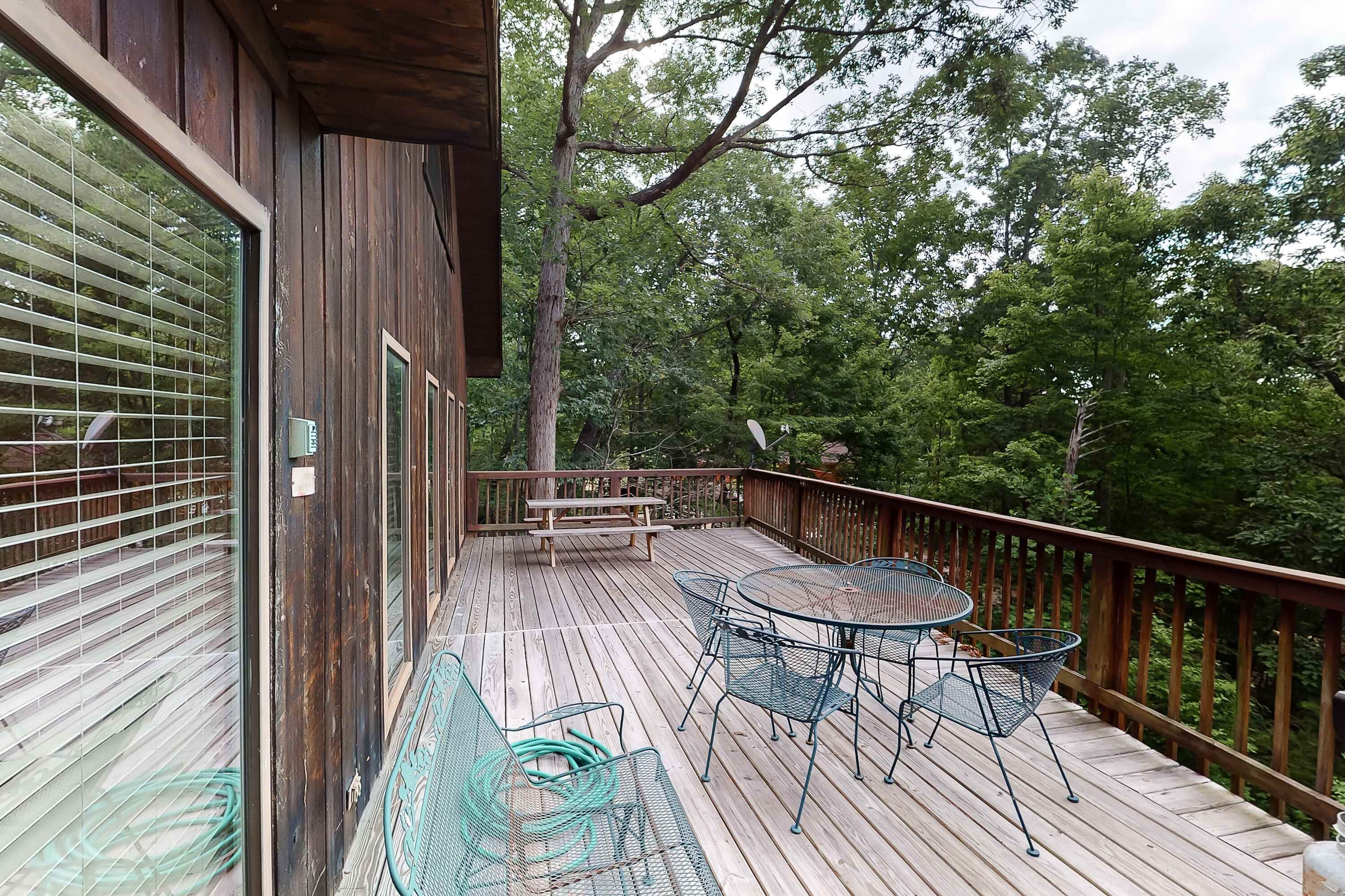 A wooden deck with a picnic table and metal chairs, surrounded by trees.