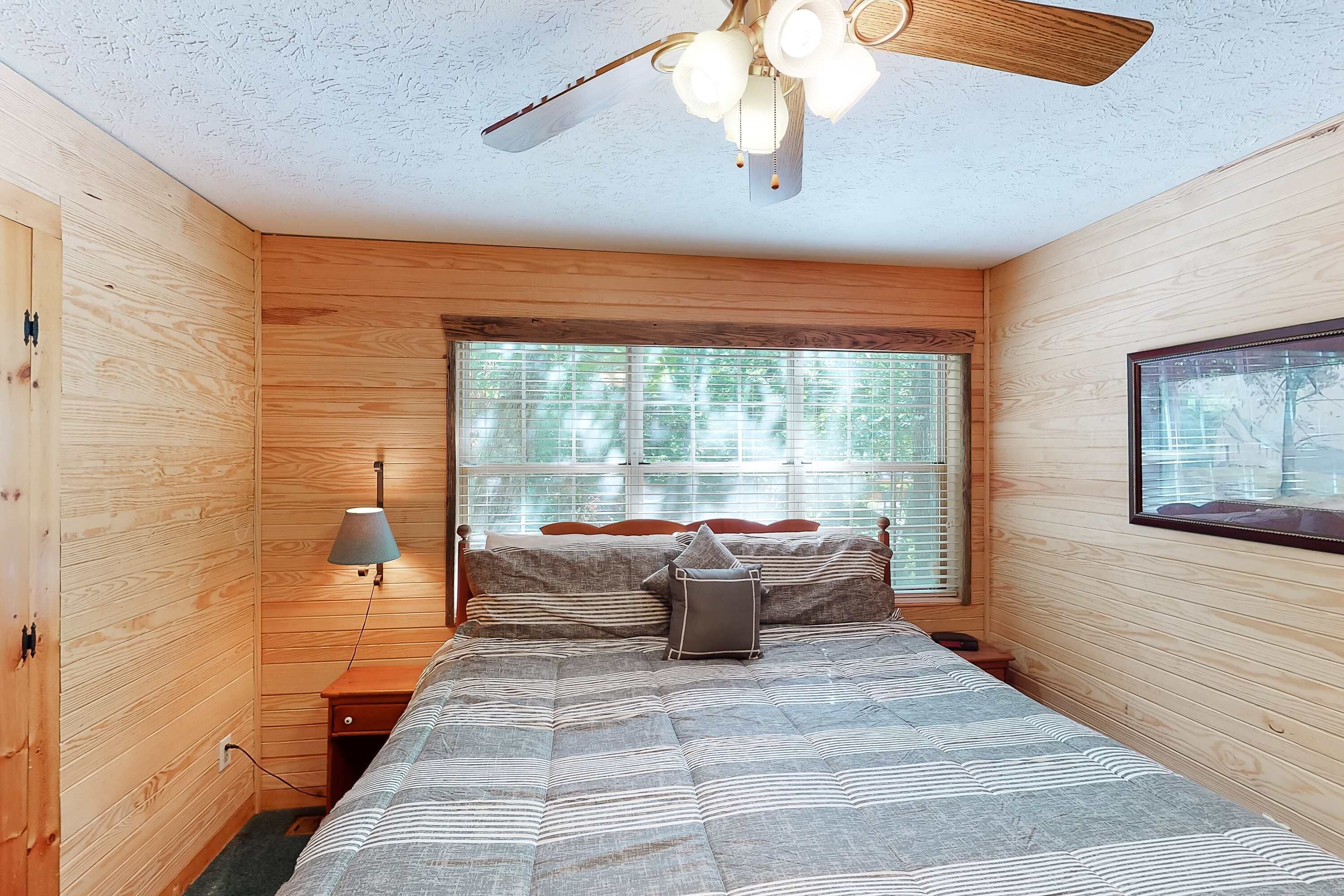 A bedroom with a bed, a nightstand, a lamp, a ceiling fan, and a window with blinds.