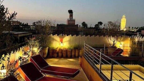 A rooftop terrace with red lounge chairs, a railing, and a view of a tower in the distance. - Riad dar Tayri (Marrakech)