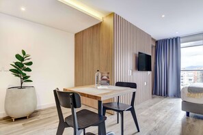Une chambre d’hôtel moderne avec une table à manger en bois, deux chaises, une plante en pot, un téléviseur à écran plat fixé au mur et une grande fenêtre avec des rideaux.