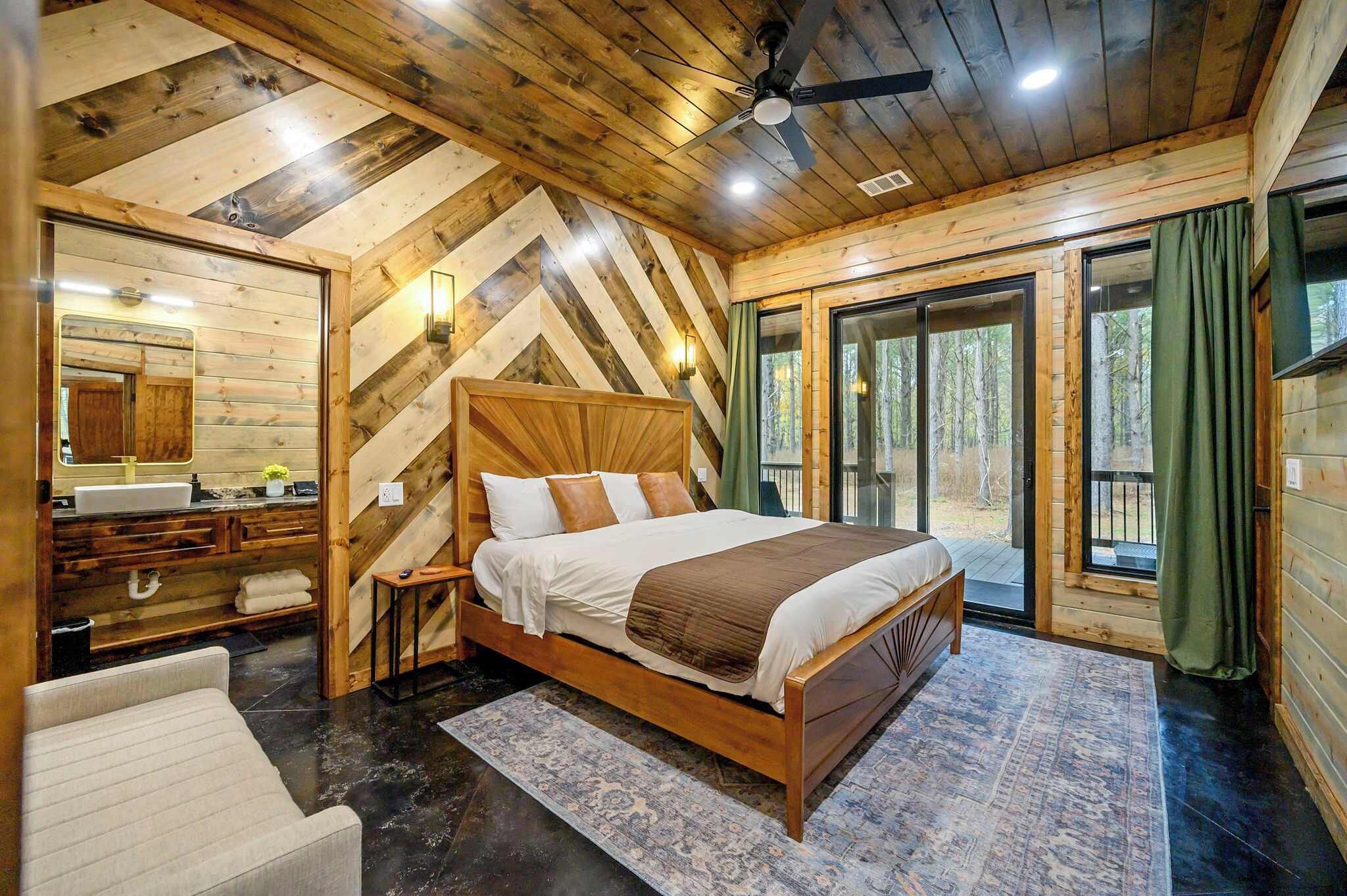 Une chambre aménagée dans une cabane en bois, avec un grand lit, une coiffeuse avec un miroir et une vue sur la forêt grâce à de grandes fenêtres.