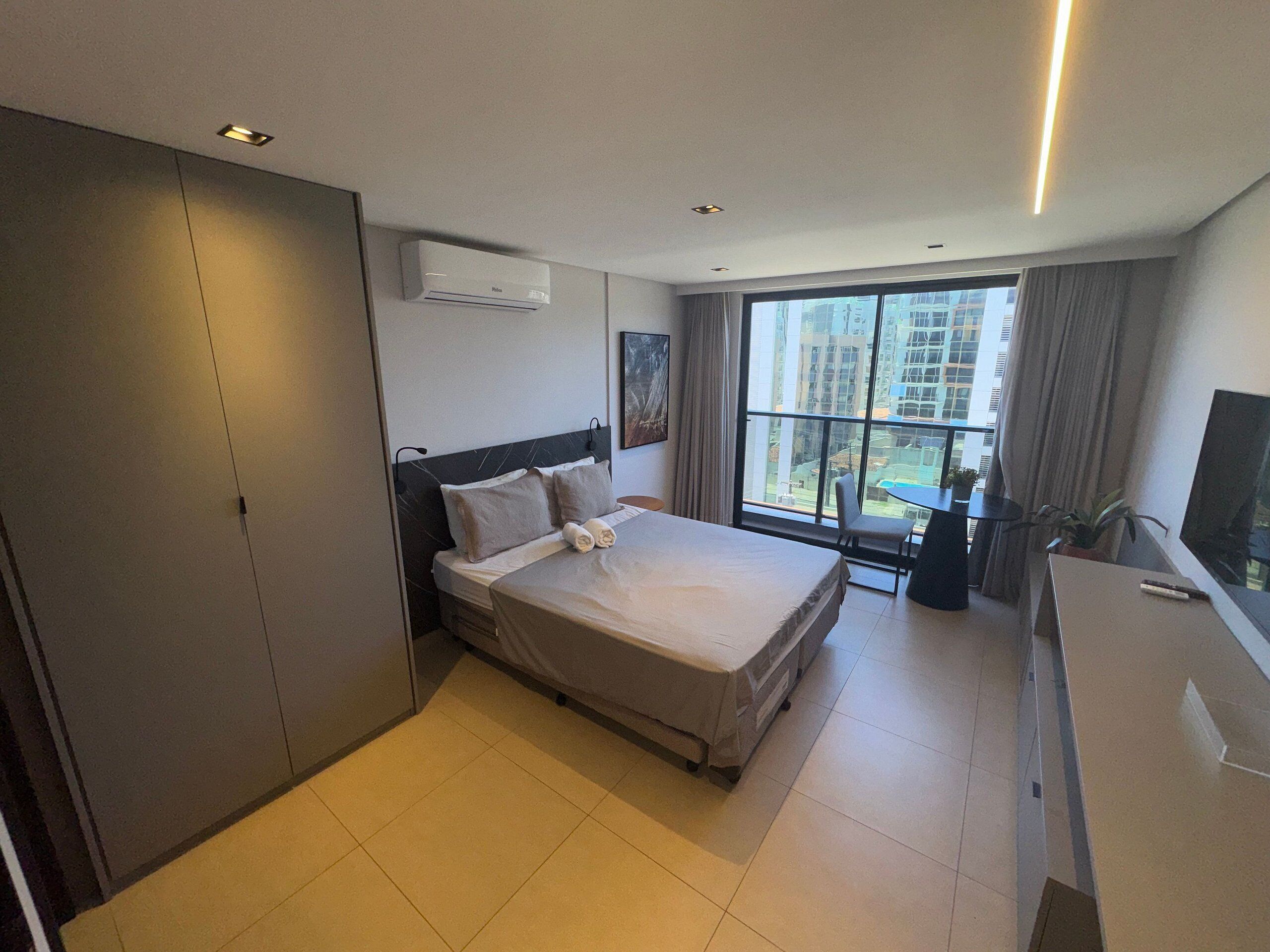 A hotel room with a bed, a desk, a TV, a window with a city view, and an air conditioning unit.