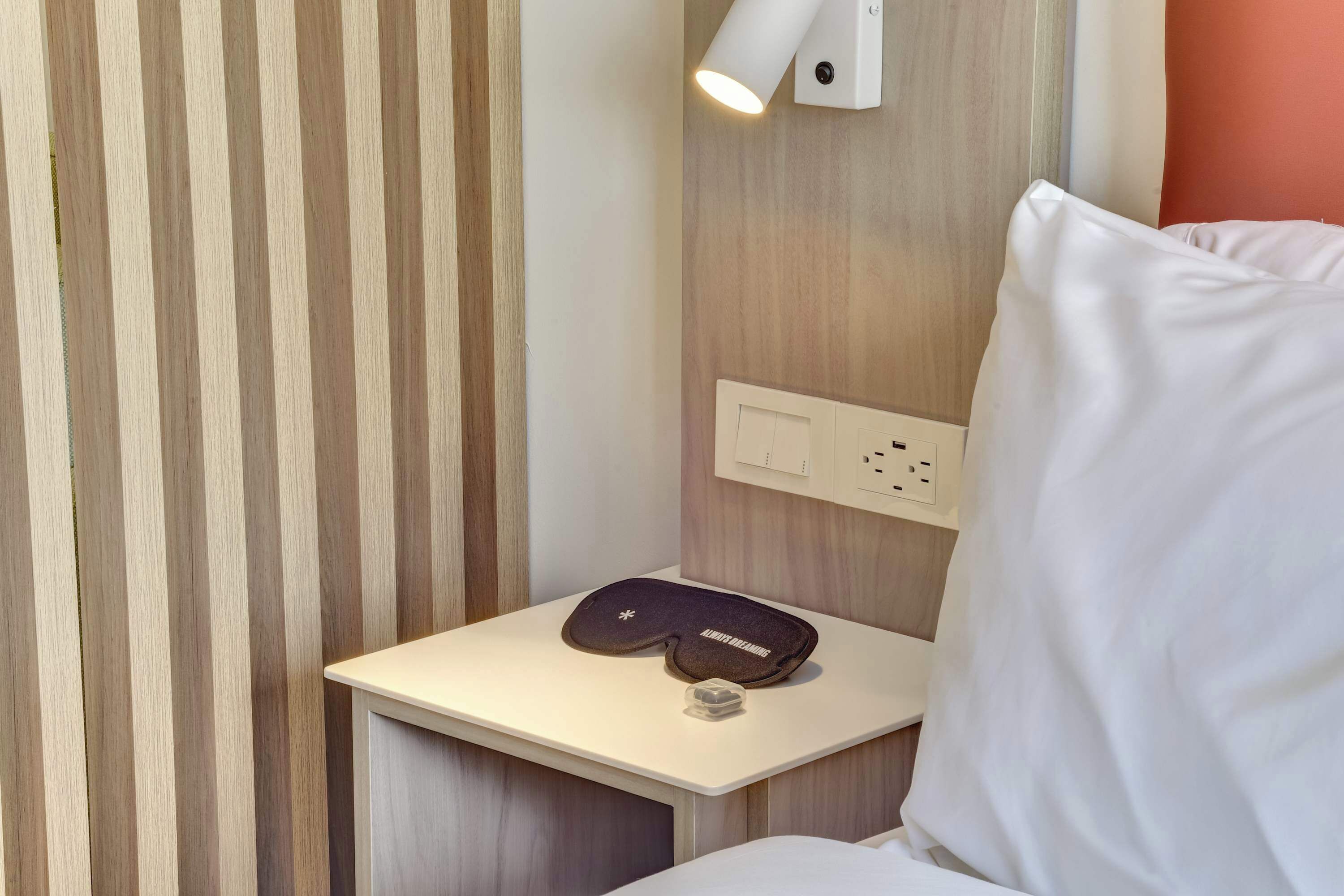 A hotel room with a striped wall, a bedside table with a pillow and a remote control, and a wall-mounted light fixture.